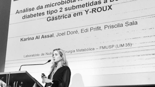 Karina apresentando a pesquisa; o slide mostra os autores Karina Al Assal e Joel Doré, do Laboratório de Nutrição e Cirurgia Metabólica da FMUSP